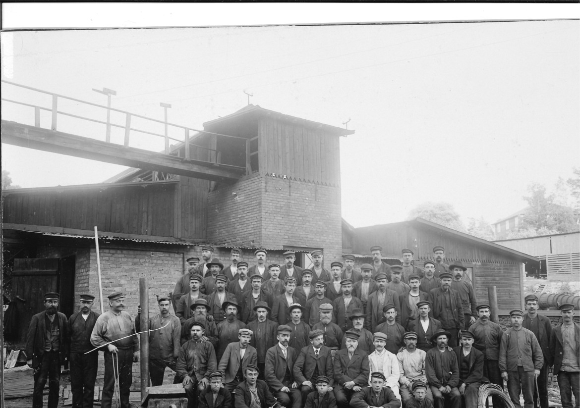 Äldre svartvitt foto med ett 60-tal uppställda arbetare framför bruksbyggnad