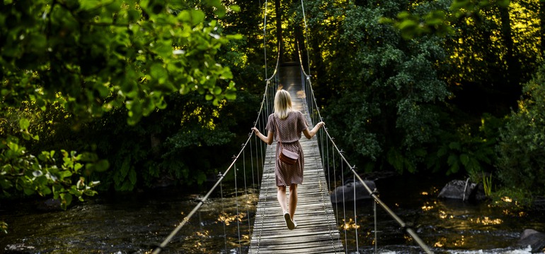 En person går över en å på en hängbro. I bakgrunden syns en grönskande skog.