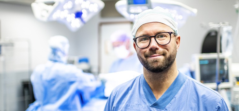 En sjuksköterska står i en operationssal. I bakgrunden pågår en operation.